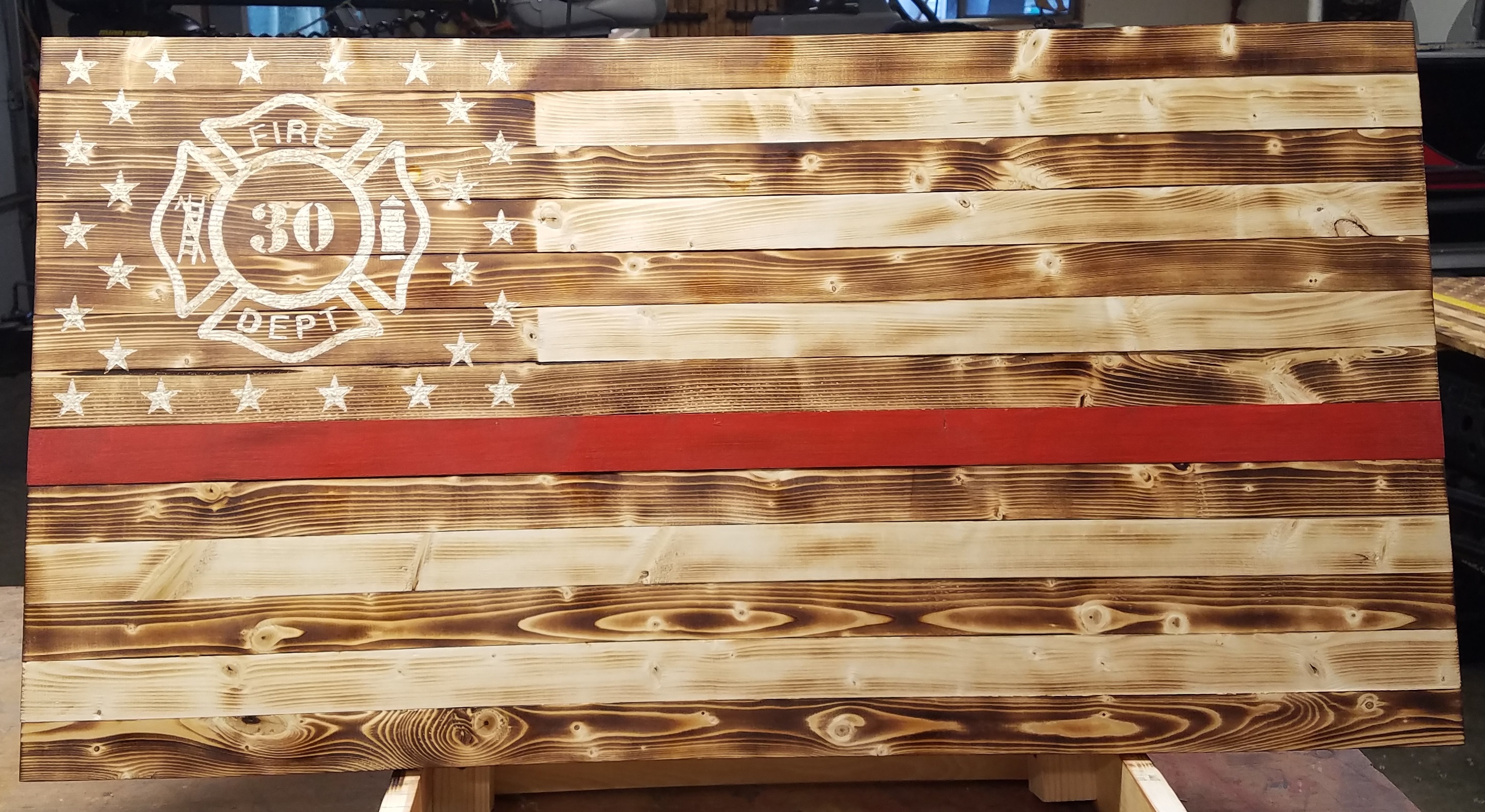 Picture of rustic fire fighter flag with thin red line painted across the middle. Located around the stars of the flag is the fire fighter symbol with customized fire figher number.